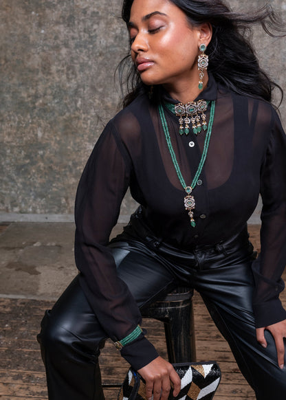 Woman in black outfit with jewelry sitting on a wooden stool against a textured wall.