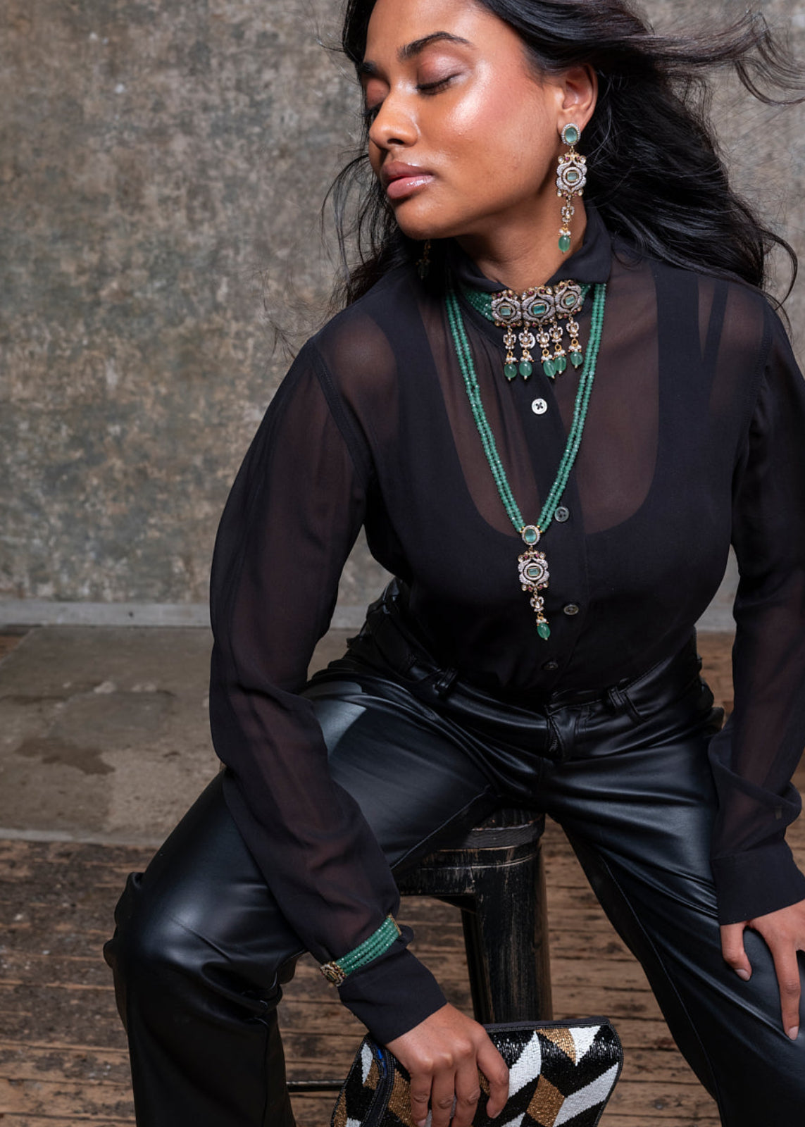 Woman in black outfit with jewelry sitting on a wooden stool against a textured wall.