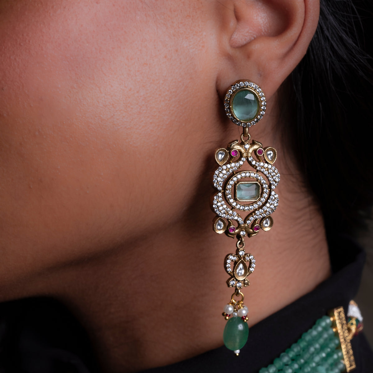 Close-up of an ornate earring with gemstones on a person wearing a black outfit with green jewelry.