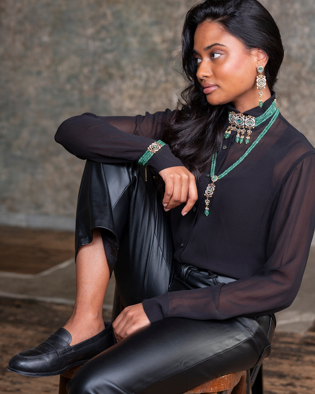 Woman sitting on a stool wearing a black outfit with jewelry against a neutral background