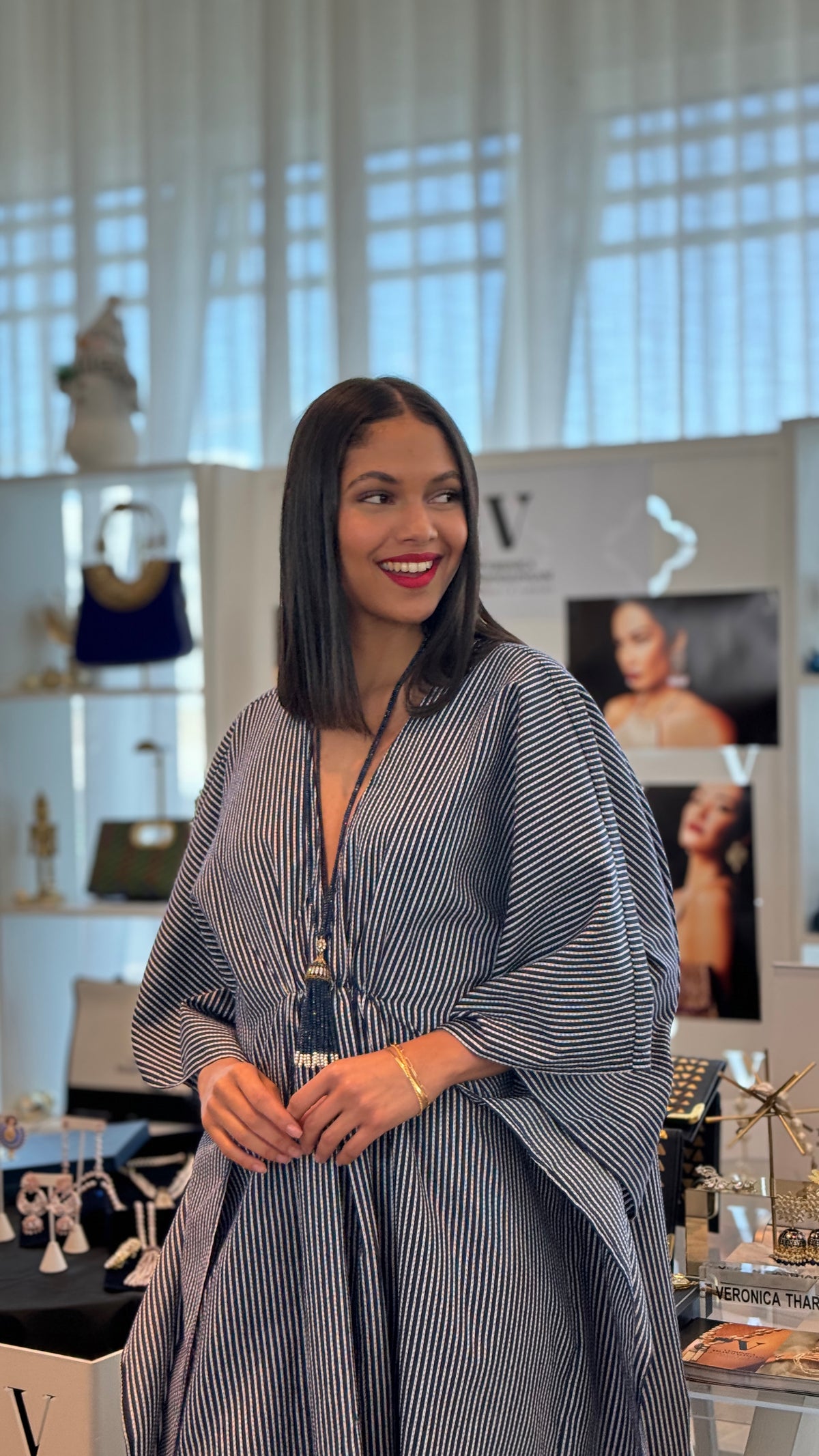 A woman with shoulder-length dark hair stands indoors wearing a loose, draped blue and white vertically striped poncho-style dress. She is smiling and looking to the side in a boutique setting with display shelves and framed photos in the background.