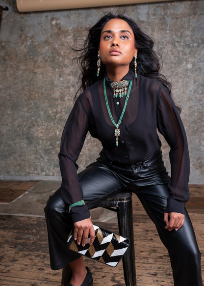 Woman in black outfit with green jewelry sitting on a stool against a textured wall.