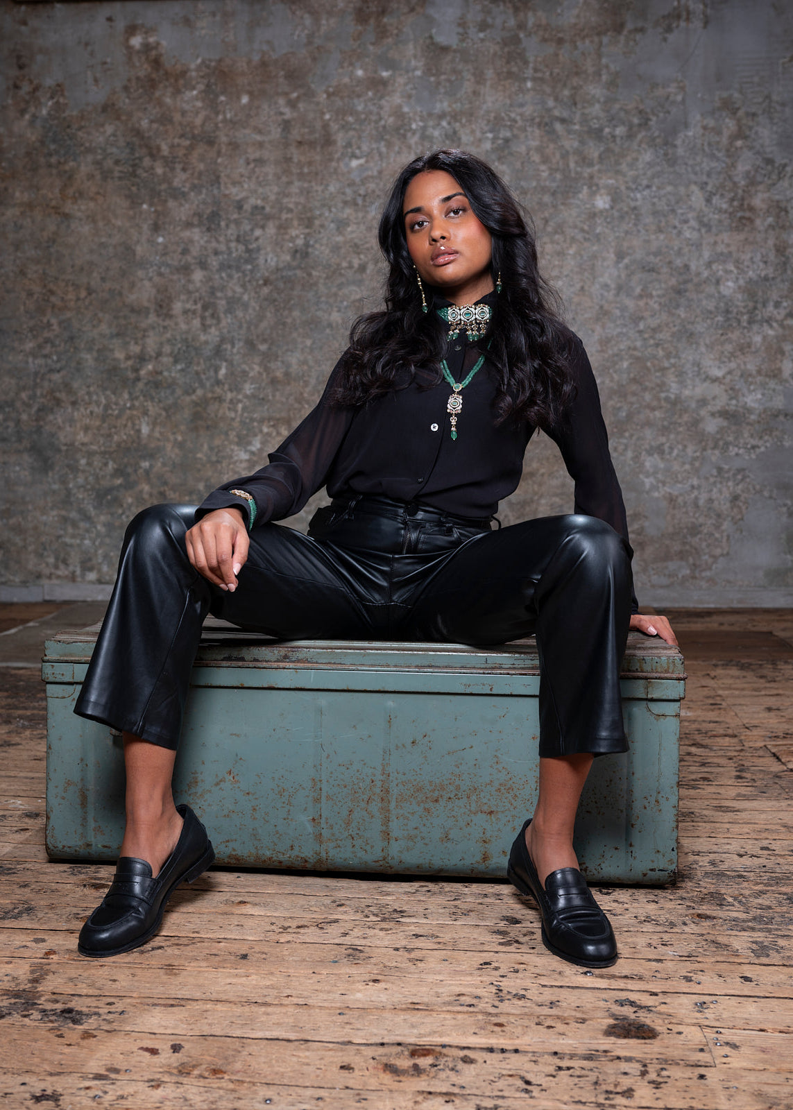 Person wearing a black outfit sitting on a rustic wooden bench against a textured wall.