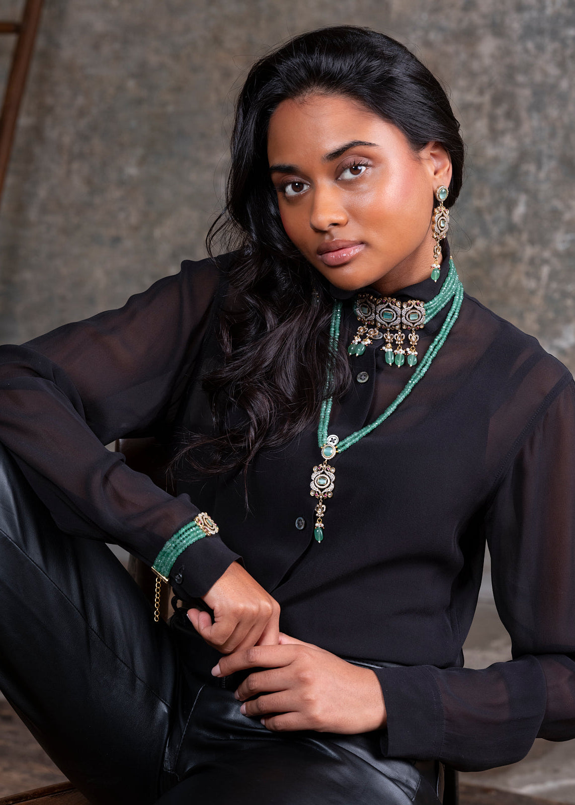 Woman wearing a black outfit with green jewelry against a textured wall.